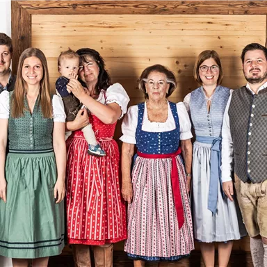 Eine Gruppe von sechs Personen in traditioneller Kleidung steht vor einer Holzwand. Sie lächeln freundlich in die Kamera.