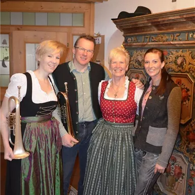 A group of four people in traditional costume stands in a cozy room. In the foreground, red roses and a decorative cabinet are visible.