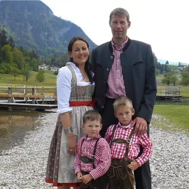 Eine Familie steht am Ufer eines Sees, umgeben von Bergen und Grünpflanzen. Alle tragen traditionelle bayerische Trachten.