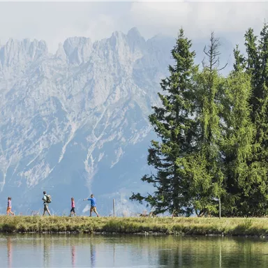 Eine Gruppe von Wanderern spaziert entlang eines ruhigen Sees, umgeben von hohen Bergen und dichten Bäumen. Die Landschaft ist malerisch und einladend.