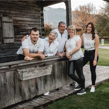 Eine Gruppe von fünf Personen steht lächelnd vor einem Holzgebäude. Sie tragen ähnliche Kleidung und wirken freundlich und einladend.