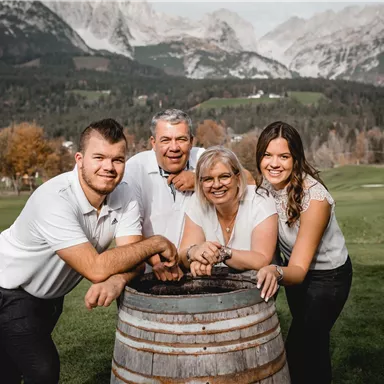 Eine Familie posiert fröhlich an einem Weinfass im Freien. Im Hintergrund sind Berge und Bäume zu sehen.