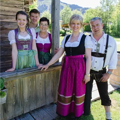 Eine fröhliche Gruppe von fünf Personen in traditioneller bayerischer Tracht steht vor einem Holzgebäude. Sie lächeln und wirken entspannt, umgeben von einer grünen Landschaft.