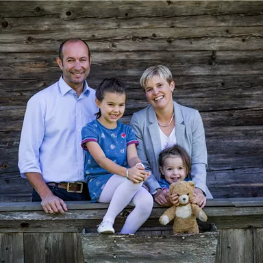 A family is standing in front of a wooden wall. Two girls are holding a teddy bear and smiling.