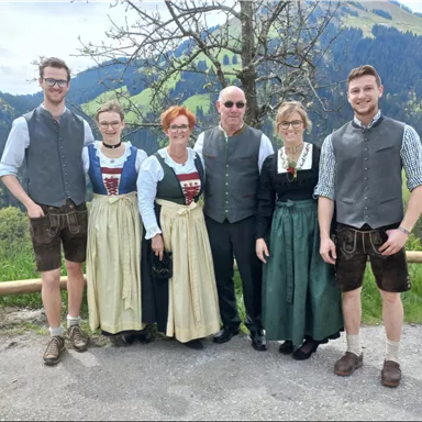 Eine Gruppe von sechs Personen in traditioneller Tracht steht vor einer malerischen Berglandschaft. Sie lächeln und posieren vor einem Hintergrund mit grünen Hügeln und Bäumen.