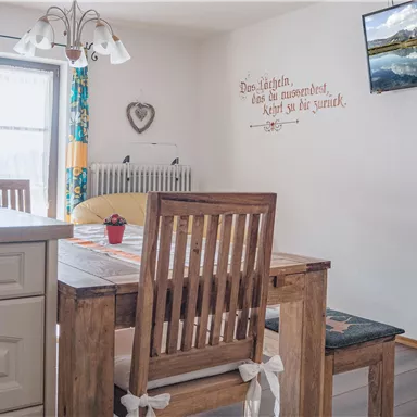 Ein gemütlicher Essbereich mit einem Holztisch und Stühlen. An der Wand hängt ein Fernseher, und das Licht fällt durch das Fenster.