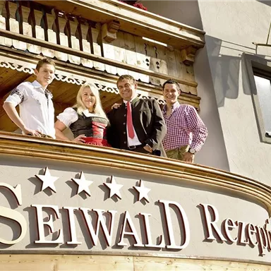 A group of four people stands on a balcony in front of a building. The sign with the name "Seiwald Rezept" is clearly visible.