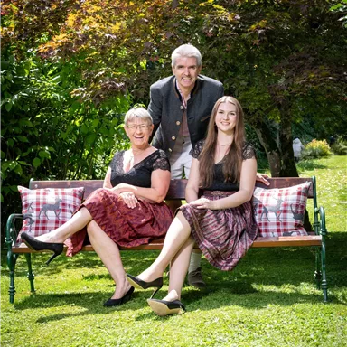 Drei Personen sitzen auf einer Bank in einem Garten. Es ist sonnig und die Umgebung ist grün und lebendig.