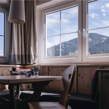 Ein gemütlicher Raum mit einem Holz-Tisch und Stühlen. Durch die Fenster sieht man die Berge und den blauen Himmel.