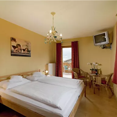 Ein gemütliches Schlafzimmer mit einem Doppelbett und warmer Farbgestaltung. Es gibt ein Fenster, einen kleinen Tisch mit Stühlen und einen Fernseher an der Wand.