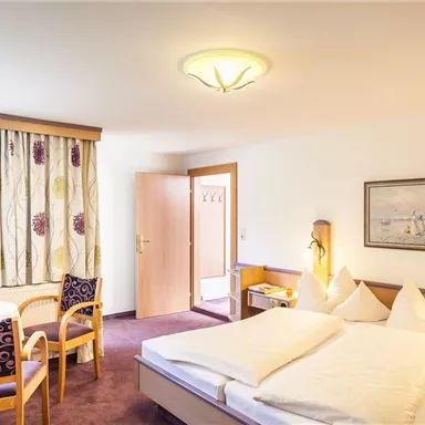 Ein gemütliches Zimmer mit einem Doppelbett und Sessel. Am Tisch stehen Getränke und es gibt einen großen Fenster mit viel Licht.