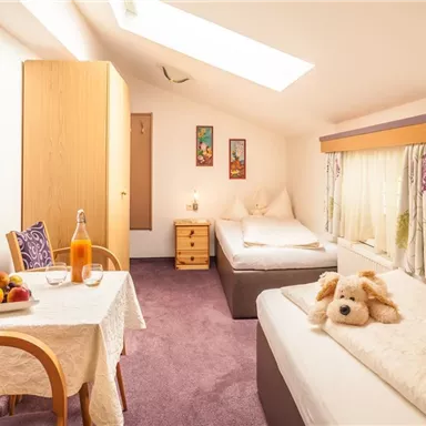 Ein gemütliches Zimmer mit zwei Betten und einem Tisch, der mit Früchten und Getränken gedeckt ist. Der Raum hat ein Dachfenster und freundliche Dekorationen.