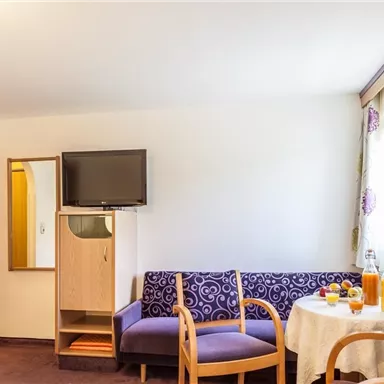 Ein gemütlicher Wohnbereich mit einem lila Sofa und einem Tisch. Im Hintergrund befindet sich ein Fernseher und ein Fenster mit Vorhängen.