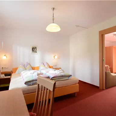 Ein helles Zimmer mit zwei Betten und einem Tisch. Der Raum hat große Fenster und einen gemütlichen Sitzbereich.