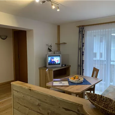 Ein gemütlicher Wohnraum mit einem Tisch, Stühlen und einem Fernseher. Es gibt große Fenster, die viel Licht hereinlassen.