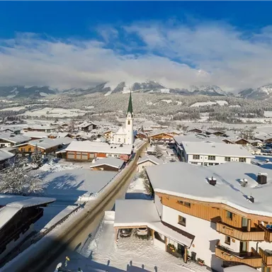 Ein malerisches Dorf im Winter mit verschneiten Hügeln und einer Kirche. Der klare blaue Himmel kontrastiert mit der weißen Landschaft.
