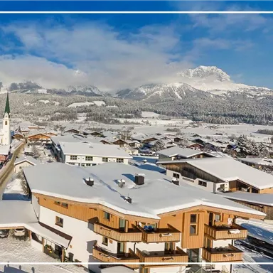 Eine malerische Winterlandschaft mit schneebedeckten Dächern und Bergen im Hintergrund. Ein Kirchenturm ragt in den klaren blauen Himmel.
