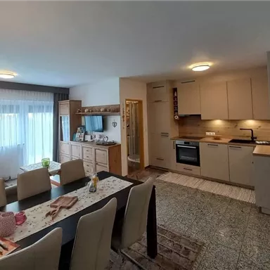 Ein modernes Wohnzimmer mit einer offenen Küche. Es gibt einen Esstisch mit Stühlen und große Fenster, die viel Licht hereinlassen.