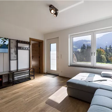 Ein helles Wohnzimmer mit großen Fenstern und Blick auf die Berge. Die Einrichtung ist modern und einladend, mit einem gemütlichen Sofa und einer Fernsehecke.