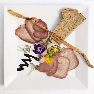 Ein eleganter Teller mit dünn geschnittenem Fleisch, verziert mit essbaren Blumen und frischen Kräutern. Dazu ein Stück dunkles Brot und eine knusprige Brotstange.