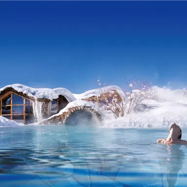 A relaxing warm water basin in a snowy landscape. In the background, snow-covered mountains and a wooden cabin can be seen.