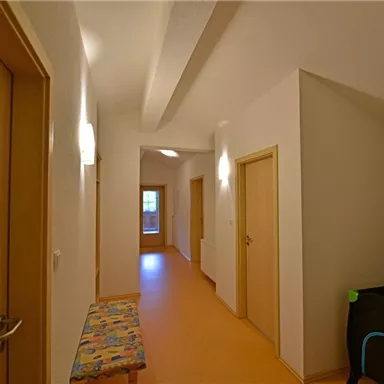 A bright hallway with light walls and wooden doors. At the end of the hallway, more doors can be seen, and a bench is positioned against the wall.