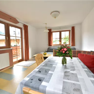 A bright and cozy living room with a table and chairs. On the table is a blooming plant, and large windows let in plenty of daylight.