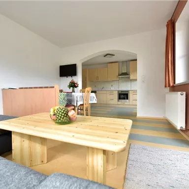 A bright living room with a modern table and a cozy couch. In the background, an open kitchen and a dining area are visible.