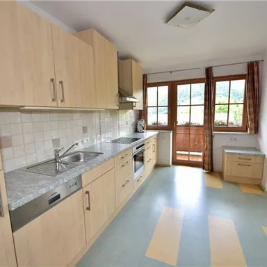 A bright, modern kitchen with wooden cabinets and a window that leads to a balcony. The floor features a decorative color scheme in blue and yellow.