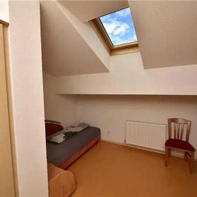 A simple room with two beds, a chair, and a heater. The window in the roof area lets in natural light.