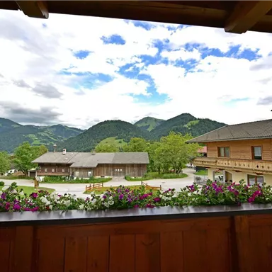 A picturesque mountain landscape with green hills and clouds in the sky. In the foreground, colorful flowers are blooming on a wooden terrace.