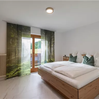 Ein modernes Schlafzimmer mit einem großen Bett und holzfarbenen Möbeln. Die großen Vorhänge zeigen ein Waldmotiv und lassen viel Licht herein.