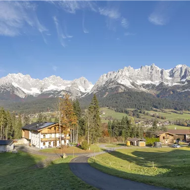 Eine malerische Landschaft mit majestätischen Bergen im Hintergrund. Im Vordergrund sind typische alpine Häuser und grüne Wiesen zu sehen.