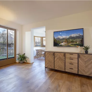 Ein modernes Wohnzimmer mit Holzfußboden und einem Fernseher an der Wand. Große Fenster bieten einen Blick ins Freie und viel Tageslicht.