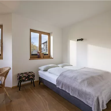 Ein helles Schlafzimmer mit zwei Fenstern und Blick auf die Berge. Es gibt ein Bett, einen Stuhl aus Rattan und einen kleinen Hocker.