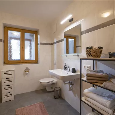 Ein modernes Badezimmer mit hellen Fliesen und einem großen Spiegel. Handtücher sind ordentlich auf einem Regal gestapelt, und es gibt ein Fenster mit natürlichem Licht.