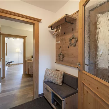 Ein heller Flur mit Holzverkleidung und einer gemütlichen Sitzbank. An der Wand sind Garderobenhaken und eine gläserne Tür, die in einen weiteren Raum führt.