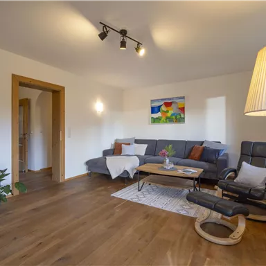 Ein gemütliches Wohnzimmer mit einer grauen Couch, einem Holzkaffee-Tisch und einer Stehlampe. An der Wand hängt ein buntes Gemälde, und der Boden ist aus Holz.