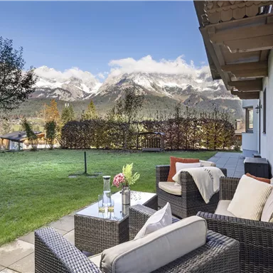 Eine gemütliche Terrasse mit Rattanmöbeln und einem Tisch im Vordergrund. Im Hintergrund sind majestätische Berge und ein klarer Himmel zu sehen.