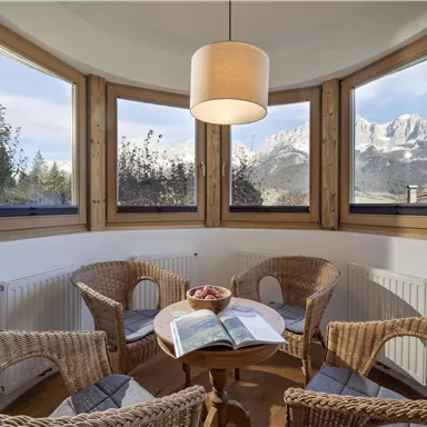 Ein gemütlicher Innenraum mit einem Tisch und vier Stühlen aus Rattan. Die großen Fenster bieten einen Blick auf die Berge und die Natur draußen.