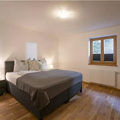 Ein gemütliches Schlafzimmer mit einem großen Bett und hellen Wänden. Das Fenster lässt viel Tageslicht herein und der Boden ist aus Holz.