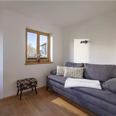 Ein gemütliches Wohnzimmer mit einem grauen Sofa und hellen Fenstern. Der Holzfußboden und die neutralen Farben schaffen eine einladende Atmosphäre.