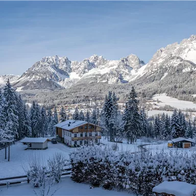 Eine verschneite Winterlandschaft mit majestätischen Bergen im Hintergrund. Im Vordergrund steht ein charmantes Holzhaus umgeben von schneebedeckten Bäumen.