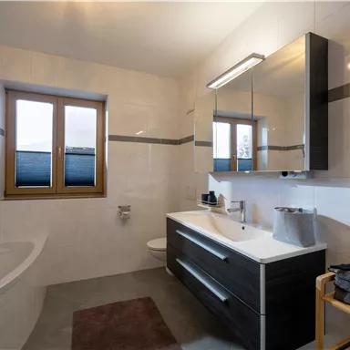 Ein modernes Badezimmer mit einem großen Spiegel und einem Waschbecken. Die Wände sind hell, und es gibt ein Fenster, das Tageslicht hereinlässt.