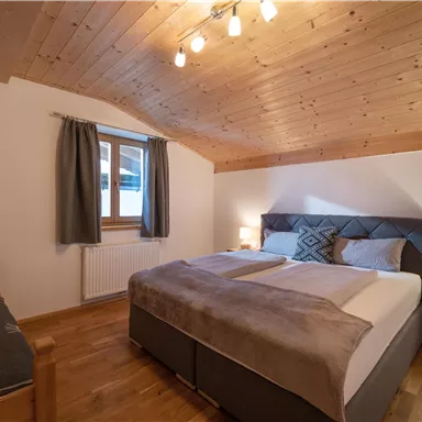 Ein gemütliches Schlafzimmer mit Holzdecke und warmem Licht. Es gibt ein großes Bett und einen bequemen Sessel in der Ecke.