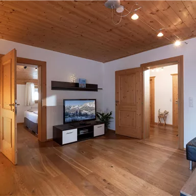 Ein schöner Wohnraum mit Holzdecke und warmen Farbtönen. Es gibt einen Fernseher, einen Couchtisch und Zugang zu weiteren Zimmern.