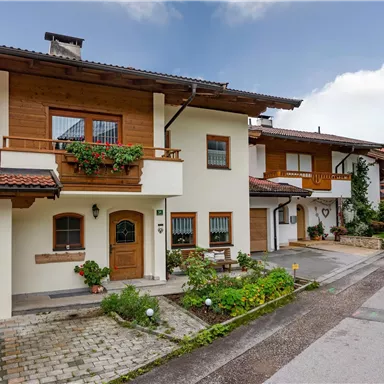 Ein charmantes Haus im alpinen Stil mit Holzverkleidung und einem begrünten Vorgarten. Die Straße ist ruhig und von Bäumen gesäumt.
