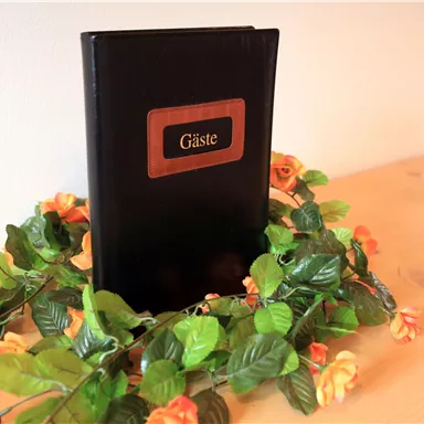Ein schwarzes Gäste-Buch liegt auf einem Tisch, umgeben von einer Dekoration aus grünen Blättern und orangen Blumen. Die Oberfläche ist aus Holz und wirkt warm und einladend.