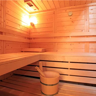 Eine gemütliche Sauna mit hölzernem Interieur und weichen Bänken. Ein Holzbehälter mit einem Löffel steht im Raum.