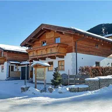 A beautiful chalet in the snow, surrounded by a clear blue sky and mountains in the background. The buildings have traditional wooden elements and an inviting atmosphere.